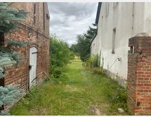 Schmaler, grasbewachsener Weg zwischen einem roten Backsteingebäude mit einer weißen Holztür links und einem weiß verputzten Gebäude rechts. Im Hintergrund sind Bäume und ein bewölkter Himmel zu sehen.