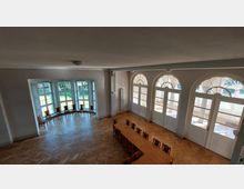 Ein heller Raum mit großen Rundbogenfenstern, die auf eine Terrasse oder Garten hinausblicken. Der Raum besitzt einen Parkettboden, einen langen Holztisch mit Stühlen und eine Sitzgruppe vor bodentiefen Fenstern an einer Seite.