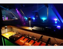Innenansicht eines Nachtclubs mit DJ-/Technikpult im Vordergrund, Mischpult und Kabeln sowie kleinen Tischlampen entlang einer niedrigen Trennwand. Im Hintergrund sind Lounge-Sitzbereiche und eine Bar zu sehen, beleuchtet von farbigen Scheinwerfern und LED-Licht in Blau, Violett und Pink unter einer schrägen Decke.