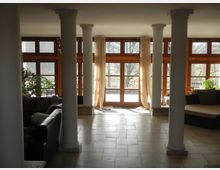 Großer, heller Innenraum mit bodentiefen Holzfenstern und cremefarbenen Vorhängen, die Tageslicht hereinlassen. Die Einrichtung umfasst weiße Säulen, Polstermöbel, gefliesten Boden und Zimmerpflanzen, mit Blick auf eine Terrasse und Bäume im Freien.