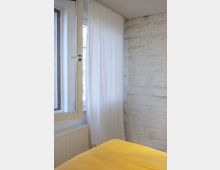 Ein Raum mit einer weißen Steinwand, einem großen Fenster mit weißem Vorhang und einer gelben Bettdecke im Vordergrund. Unter dem Fenster befindet sich ein Heizkörper.