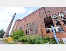Backsteinfassade eines verlassenen Fabrikgebäudes mit zerbrochenen Fenstern, einem hohen Schornstein und wuchernder Vegetation im Vordergrund. Rechts ist ein blaues Rohrsystem an der Wand befestigt, und ein gelber Baucontainer steht links im Bild.