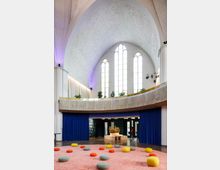 Heller Innenraum einer umgebauten Kirche mit hoher, gewölbter Decke und großen Spitzbogenfenstern. Der Raum ist modern eingerichtet mit einem rosafarbenen Teppich, bunten Sitzkissen und einer Galerieebene mit Pflanzen sowie blauen Vorhängen.