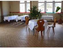 Ein heller Raum mit mehreren Tischen, die mit weißen Tischdecken bedeckt sind, umgeben von Holzstühlen mit herzförmigen Ausschnitten in den Lehnen. Fenster an der Wand lassen Tageslicht herein, Zimmerpflanzen stehen in Töpfen, und einfache Dekorationen aus Blumenvasen befinden sich auf den Tischen.