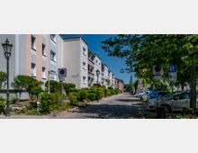 Wohnstraße mit mehrstöckigen Wohnhäusern auf der linken Seite und geparkten Autos auf der rechten Seite entlang eines Kopfsteinpflasterwegs. Straßenlaternen, Verkehrsschilder und Bäume säumen die Straße, die von sonnigem Wetter erleuchtet ist.