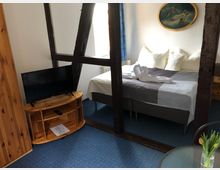 Hotelzimmer mit rustikalem Holzgebälk, einem Doppelbett mit weißer und grauer Bettwäsche, sowie gefalteten Handtüchern in Schwanenform. Im Raum befinden sich ein kleiner Fernseher auf einem Holzmöbel, ein blauer Teppichboden und ein runder Glastisch mit einer Blumenvase.