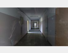 Langer, schmaler Flur in einem vermutlich verlassenen Gebäude mit kahlen, teils beschädigten Wänden und sichtbaren Türen auf beiden Seiten; der Boden ist mit quadratischen Fliesen ausgelegt, und am Ende des Flurs scheint Tageslicht herein.