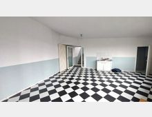 Leerer Raum mit schwarz-weiß kariertem Boden, weißen Wänden mit hellblauer unteren Hälfte, einer kleinen Spüle in der Ecke und einer offenen Tür, die zu einer Treppe führt. Rechts im Bild befindet sich eine weitere offene Tür.