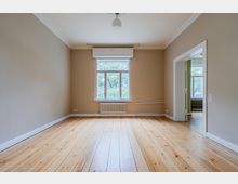 Leerer Wohnraum mit hellem Holzdielenboden, beigefarbenen Wänden und weißer Stuckleiste an der Decke. Mittig ein großes Fenster mit Heizkörper darunter und rechts eine offene Tür zu einem angrenzenden Zimmer.