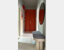 Narrow apartment entry hallway with light gray walls and white tiled floor, leading to a large red double door with matching built-in cabinets above. A round wall mirror and a cushioned bench sit along the right wall, with a framed poster on the left and doorways opening off the corridor.