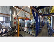 Industriehalle mit Rohrleitungen und Ventilatoren. Zu sehen sind gelbe und silberne Rohre, blaue Stahlträger sowie Belüftungsanlagen, umgeben von Laufstegen mit Geländern, unter einem Metalldach mit sichtbaren Balkenkonstruktionen.