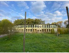 Ein modernes, zweistöckiges Gebäude mit großen Fenstern und hellgrüner Fassade, umgeben von einer grünen Wiese und Bäumen. Der Himmel ist leicht bewölkt, und im Vordergrund stehen vereinzelt Holzpfähle.
