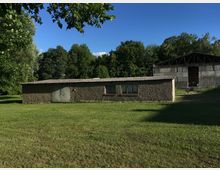 Flache, graue Gebäude mit Wellblechdächern in einer ländlichen Umgebung, umgeben von einer großen Wiese und hohen Bäumen im Hintergrund. Im Vordergrund ist ein kleineres Gebäude mit Metalltüren und schmalen Fenstern, während rechts ein größeres, halboffenes Gebäude zu sehen ist.