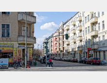 Straßenszene mit mehrstöckigen Wohnhäusern und einem Bistro im Eckgebäude links, daneben geparkte Autos und Fahrräder. Im Hintergrund ist leicht versetzt der Berliner Fernsehturm erkennbar, während eine Ampel den Übergang markiert.