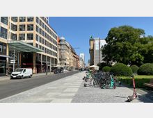 Straßenszene in einer urbanen Umgebung mit modernen und historischen Gebäuden; links ein Einkaufszentrum mit Glasüberdachung, daneben ein älteres Gebäude mit Kuppeldach. Rechts ein Gehweg mit geparkten Fahrrädern und E-Scootern sowie Grünanlagen mit Bäumen und Sträuchern.