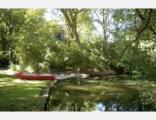 Ein kleiner Teich in einem Waldgebiet mit dichtem Baumbewuchs. Am Ufer liegt ein rotes Ruderboot auf einer Grünfläche, und das Wasser reflektiert die umgebenden Bäume.