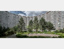 Ein großer, halbkreisförmiger Wohnblock mit vielen Balkonen und Fenstern umgibt eine begrünte Außenfläche mit Rasen, Bäumen und einem zentralen gepflasterten Bereich. Der Himmel ist bewölkt, und die Szenerie wirkt von Pflanzen und Gehwegen geprägt.