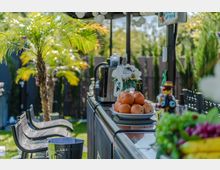 Ein offener Außenbereich mit einer Theke, auf der ein Teller mit Orangen, Getränkeflaschen und Dekorationsgegenständen steht. Im Hintergrund sind Palmen, Rasen und Holzzäune zu sehen.