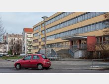 Gelbes, rechteckiges Gebäude mit mehreren Etagen, großen Fensterreihen und einem überdachten Eingangsbereich mit Treppe; im Vordergrund ein rotes Auto auf einer Straße und ein Gehweg mit Laternenmast. Im Hintergrund sind Wohnhäuser und kahle Bäume zu sehen.