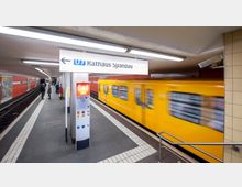 U-Bahn-Station mit gelbem Zug auf der Linie U7 in Richtung „Rathaus Spandau“. Die Station hat rote Wandverkleidungen, eine SOS-Säule und ein Schild mit Linien- und Zielangabe, während Personen auf dem Bahnsteig warten.