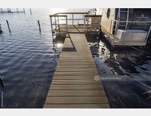 Steg aus Holzplanken, der über das Wasser zu einer Plattform führt, neben der ein Hausboot mit metallischen Details angedockt ist. Im Hintergrund spiegeln sich Sonnenstrahlen auf der ruhigen Wasseroberfläche.