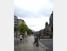 Straßenansicht in einer städtischen Umgebung mit Gehweg, niedrigen Bäumen und Sitzbereichen auf der linken Seite, flankiert von modernen Gebäuden. Rechts im Bild ist ein historisches Bauwerk mit Kuppel und Steinfassade zu sehen, im Hintergrund erstreckt sich die Skyline mit weiteren Gebäuden.