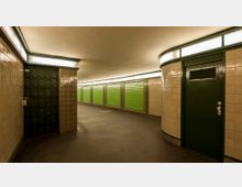 Unterirdischer Gang mit hellbraunen, gefliesten Wänden, grünen Türen und grünen Wandpaneelen. Die Beleuchtung erfolgt durch lange Leuchtbänder an der Decke.