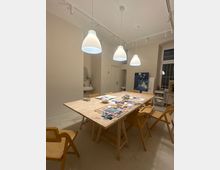 Heller Atelier- oder Workshopraum mit großem Holztisch in der Mitte, umgeben von mehreren Holzstühlen und mit Papier, Farben und Bastelmaterialien auf dem Tisch. Über dem Tisch hängen drei weiße Pendelleuchten; im Hintergrund stehen Regale und ein großes Gemälde an der Wand neben einem hohen Fenster.