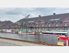 Backsteingebäude eines Hafens mit Satteldächern, davor liegt ein Wasserbecken mit mehreren Passagierschiffen und einem alten Hafenkran. Im Vordergrund sind Pflastersteine und Schienen sichtbar.