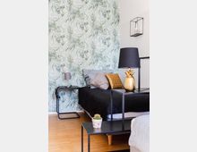 Schlafzimmer mit modernem Design, einer Tapete mit tropischem Blattmuster an der Wand, einem Bett mit schwarzer Bettwäsche und dekorativen Kissen. Daneben steht ein Metall-Nachttisch mit Lampe; im Vordergrund ein kleiner Kaktus auf einem Beistelltisch.