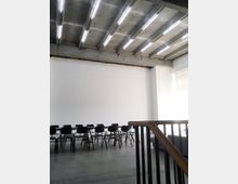 Großer, minimalistisch gestalteter Raum mit einer Betondecke und Reihen aus rechteckigen Deckenleuchten. Im Vordergrund ein Geländer, dahinter eine lange Tischreihe mit schwarzen Stühlen vor einer weißen Wand.