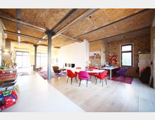 Großzügiger Wohn- und Arbeitsbereich in einem Loft mit Backsteinwänden, freigelegten Stahlträgern und einer gewölbten Ziegeldecke. Der Raum ist mit einer offenen Küche, einem Esstisch mit bunten Stühlen, einem Schreibtisch und mehreren Sitzgelegenheiten eingerichtet; große Fenster lassen viel Tageslicht herein.