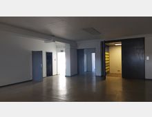 Ein leerer, industrieller Innenraum mit grauem Betonboden und weiß gestrichenen Wänden. Zu sehen sind mehrere geöffnete Türen, darunter eine schwarze Schiebetür zu einem Aufzug mit gelb-schwarzem Streifenmuster im Inneren, sowie ein Notausgang mit Tageslichteinfall.