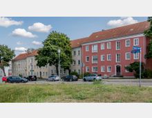 Wohngebäude in einer urbanen Umgebung mit hellbeigen und rostroten Fassaden, drei Stockwerken und weißen Sprossenfenstern. Im Vordergrund eine Straße mit parkenden Autos, einer Straßenlaterne, einem Straßenschild und grasbewachsenen Grünflächen, daneben stehen Bäume.