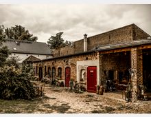 Ein rustikaler Hof mit Backsteingebäuden, roten Türen und gewölbten Fensteröffnungen, umgeben von Pflanzen und dekorativen Elementen. Im Vordergrund befinden sich Gartenmöbel auf einem gepflasterten Boden, während im Hintergrund ein weiteres Haus mit grauem Ziegeldach sichtbar ist.