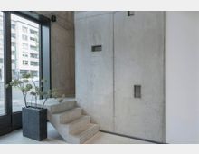 Innenraum mit modernem, minimalistischem Design: Eine rohe Betonwand mit kleinen rechteckigen Öffnungen, daneben eine schmale Treppe aus Beton. Links steht ein rechteckiger Pflanzkübel mit einer Zimmerpflanze vor einer großen Glasfensterfront, die den Blick auf eine Stadtstraße freigibt.