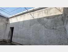 Außenhof mit hohen, grauen Betonwänden, einer kleinen Türöffnung und einem Drahtgitter, das den oberen Bereich abdeckt. Darüber befindet sich ein Geländer, und der Himmel ist teilweise sichtbar.