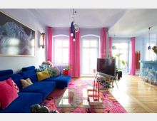 Bright, modern living room with a blue sectional sofa and colorful pillows, a glass coffee table on a patterned pink rug, and a TV on a stand. Large white-framed windows and a glass door are framed by pink curtains, with pendant lights and an open area leading toward a kitchen or dining space.