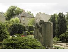 Friedhof mit einer trauernden Frauenfigur aus Stein, die sich auf einen Grabstein lehnt, umgeben von gepflegten Büschen, Bäumen und Blumenbeeten; im Hintergrund stehen zwei Wohngebäude.