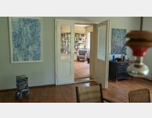 Ein Wohnzimmer mit Holzboden und weiß gestrichenen Wänden, das durch eine Glastür in einen angrenzenden Raum mit Bücherregalen führt. Im Vordergrund sind ein gemalter Wanddruck, ein Stuhl mit geflochtenem Sitz und eine alte Kommode mit Dekorationen sichtbar.