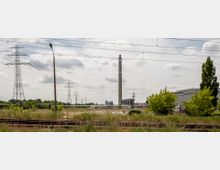 Weitläufiges Industriegebiet mit hohen Strommasten, einem Schornstein im Zentrum und Gleisen im Vordergrund. Im Hintergrund sind Büsche, niedrige Gebäude und eine teils bewölkte Himmelsszenerie zu sehen.