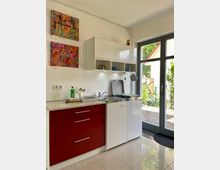 Minimalistische Küchenzeile in einem hellen Raum mit roten Unterschränken, weißen Hängeregalen und einer kleinen Edelstahlspüle. Ein großes Fenster mit Glastür bietet Blick nach draußen auf grüne Pflanzen und ein Hausdach; bunte Kunstwerke hängen an der weißen Wand.