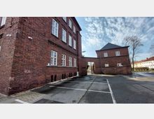 Backstein-Gebäudekomplex mit großen weißen Fenstern, verbunden durch einen Torbogen; im Vordergrund ein leerer Parkplatz mit markierten Stellflächen, im Hintergrund offener Himmel und einige Bäume.