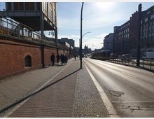 Straßenansicht mit einer erhöhten Stadtbahn, die auf einer gemauerten Brücke links fährt, und einem breiten Bürgersteig daneben. Rechts verläuft eine mehrspurige Straße mit einer gelben Straßenbahn und vereinzeltem Verkehr, flankiert von mehrstöckigen Gebäuden.