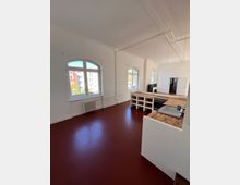 Heller, leerer Raum mit hohen Decken, weißen Wänden und großen, bogenförmigen Fenstern, die Tageslicht hereinlassen. Im Vordergrund ist eine offene Küche mit Holzarbeitsplatten und integrierten Regalfächern sichtbar; der Boden ist dunkelrot.