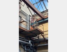 Blick in einen industriellen Innenhof mit Stahlstegen und Treppen, freiliegenden Rohrleitungen und Backstein- sowie Betonwänden. Oben überspannt ein Metallrahmen mit Drahtgitter das offene Dach und lässt den Himmel sichtbar.