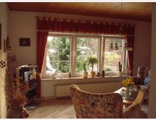 Gemütliches Wohnzimmer mit großen Fenstern und roten Vorhängen, die den Blick auf einen Garten gewähren. Im Raum befinden sich ein Fernseher, ein Blumenstrauß auf einem Glastisch, gepolsterte Sessel und mehrere Zimmerpflanzen auf der Fensterbank.