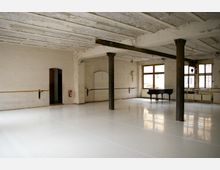 Großer, heller Tanz- oder Proberaum mit weißem Boden, weißen Backsteinwänden und einer offenen Decke mit sichtbaren Holzbalken. Der Raum verfügt über Ballettstangen, eine Feuerschutztür, einen schwarzen Flügel und große Fenster, die viel Tageslicht einlassen.