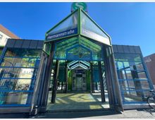 Außenansicht des gläsernen Eingangsbereichs der S-Bahn-Station Hermannstraße in Berlin, mit grünem und blauem Metallrahmen sowie reflektierenden Glasflächen; im Hintergrund sind umliegende Gebäude und klare blauer Himmel sichtbar.