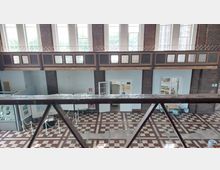 Innenansicht eines historischen Gebäudes mit roten Backsteinwänden, großen Fensterreihen und einer Galerie im oberen Stockwerk. Der Boden ist mit einem Schachbrettmuster aus roten und cremefarbenen Fliesen gestaltet, und im Vordergrund ragt ein Metallgeländer ins Bild.